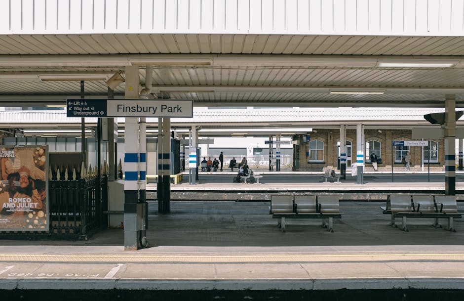 The image depicts Finsbury Park train station platform under a large, white, slatted roof with several grey metal benches arranged along the edge, providing seating for waiting passengers. A sign indicating 'Finsbury Park' is suspended from the roof structure, with additional signage directing to lifts and underground access. On the platform, there are a few individuals seated, with some standing or walking nearby. To the left, there is a black railing enclosing an advertisement board displaying promotional material, and the platform surface is made of grey concrete with tactile paving near the edge for safety. In the background, multiple train tracks extend across the width of the image, with a distant view of an old brick building and platform structures. The scene has natural daylight, illuminating the area evenly. This setting is associated with house removals or moving preparations, where a moving service such as Man with Van Honor Oak might facilitate a home relocation involving furniture transport and packing logistics at a nearby station.