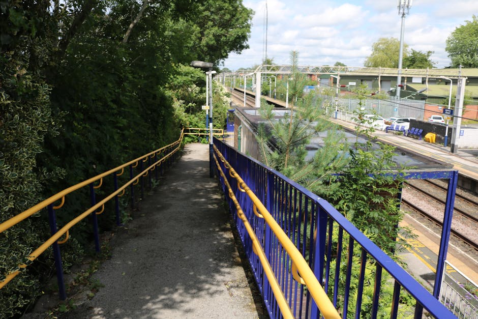 A narrow outdoor footpath with a grey, textured surface, flanked by blue metal railings with yellow horizontal handrails on both sides. The path is shaded by overhanging leafy green trees on the left, casting dappled shadows on the ground. In the distance, there is a tall, slim streetlamp positioned towards the middle of the path. The path runs parallel to railway tracks visible below and to the right, with a small cluster of trees and bushes growing alongside the rails. Across the railway, there is a station platform with a yellow safety line, a row of parked cars, and station signage. Overhead power lines and supporting structures are visible above the railway. The scene appears to be part of a transportation or home relocation environment, with a focus on the pathway leading towards the station area, possibly used by movers or residents during a house move. The image captures a clear, sunny day with a partly cloudy sky and natural lighting highlighting the different elements in the scene.