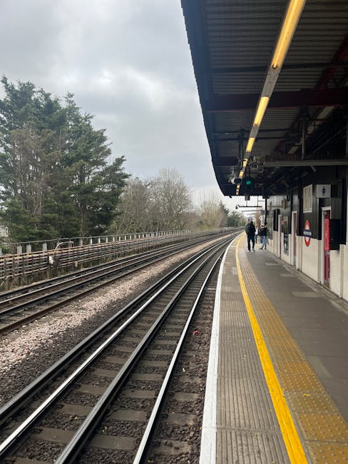 The image depicts Finsbury Park train station platform under a large, white, slatted roof with several grey metal benches arranged along the edge, providing seating for waiting passengers. A sign indicating 'Finsbury Park' is suspended from the roof structure, with additional signage directing to lifts and underground access. On the platform, there are a few individuals seated, with some standing or walking nearby. To the left, there is a black railing enclosing an advertisement board displaying promotional material, and the platform surface is made of grey concrete with tactile paving near the edge for safety. In the background, multiple train tracks extend across the width of the image, with a distant view of an old brick building and platform structures. The scene has natural daylight, illuminating the area evenly. This setting is associated with house removals or moving preparations, where a moving service such as Man with Van Honor Oak might facilitate a home relocation involving furniture transport and packing logistics at a nearby station.