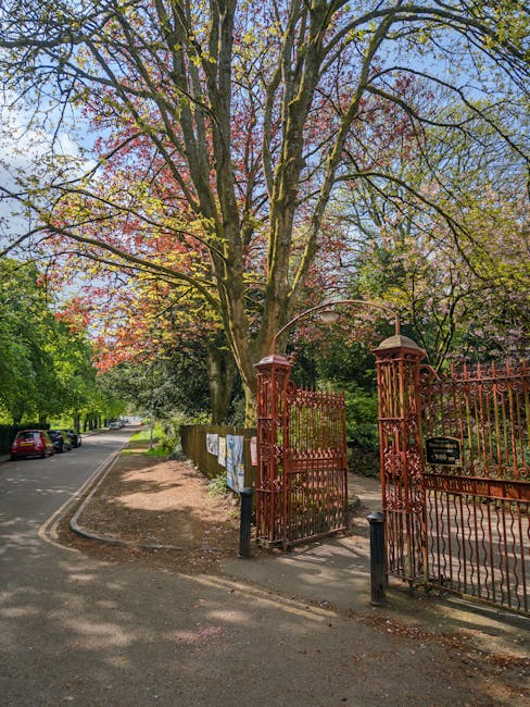 A large deciduous tree with a thick trunk and extensive branches, covered in pink and green foliage, stands behind an ornate, rust-colored wrought-iron gate with decorative spearhead finials positioned at the entrance to a property in the Horniman Museum area. The gate is slightly open, revealing a pathway leading into the garden. To the left, a narrow street with parked cars along the curb is visible, bordered by a sidewalk with some laundry hanging on a line attached to a wooden post. The scene is bathed in natural daylight, with shadows cast by the tree and fence, illustrating a peaceful urban environment suitable for house removals or property moves, with [COMPANY_NAME] providing professional transport and packing services for home relocation projects.
