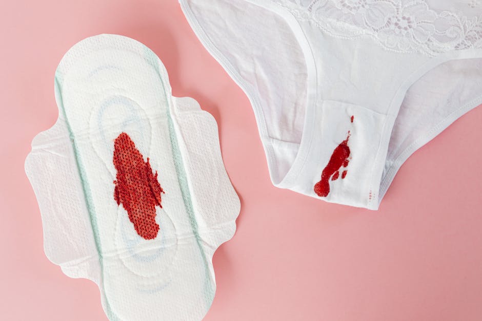 A torn disposable adult pad with red bloodstains laid flat on a light pink surface, alongside a white fabric underwear with a small bloodstain near the waistband. The image depicts a bedding or sanitary products related to personal hygiene, relevant in the context of home relocation or packing during a move. The setting appears to be indoors with soft lighting, and the objects are arranged to highlight the state of used sanitary items, which might be packed or discarded as part of packing and moving preparations by Man with Van Honor Oak in their removals service category.