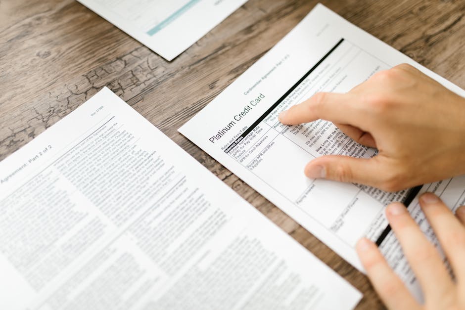 A close-up view shows a person's right hand pointing to a financial document titled 'Platinum Credit Card' that is placed on a wooden table alongside other sheets of paper containing text and data. The table surface has a natural, textured appearance with visible grain patterns and cracks. In the background, there are additional sheets of paper, possibly receipts or invoices, slightly out of focus. The image captures the process of reviewing paperwork related to home relocation expenses or moving estimates, with attention to detailed financial documentation. Occasionally, the presence of packing or moving-related context can be inferred from the nature of documents involved in an organized furniture transport or packing and moving process. This image aligns with content about avoiding hidden removal fees, which Man with Van Honor Oak provides as part of their removals services around SE23, HONOR OAK.