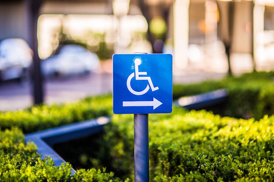 A blue parking sign indicating accessible parking with a white wheelchair symbol and an arrow pointing to the right, mounted on a metal pole in a landscaped outdoor area. The background shows blurred cars parked in the street, green bushes, and hints of buildings with warm lighting at dusk. The image highlights the designated accessible parking space outside a property, relevant to house removals and relocation services, with the sign situated on a paved pavement next to well-maintained greenery. The natural lighting and environment suggest a residential or urban setting, supporting the context of assisting clients with accessible parking when planning a home relocation or furniture transport by Man with Van Honor Oak.