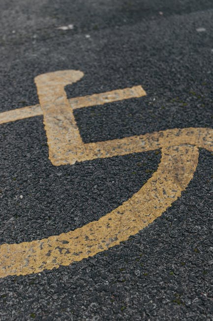 A close-up view of a yellow disabled parking bay symbol painted on black asphalt pavement, with visible texture and small cracks in the surface. The painted lines are slightly worn, with some peeling and fading. The surrounding area appears to be part of an outdoor parking space, possibly near a residential or commercial property. This image relates to house removals and relocation services offered by Man with Van Honor Oak, as it highlights parking considerations essential for planning a home relocation or furniture transport. Proper access and parking availability are critical elements during the loading process of household items, appliances, and furniture into a van, typically carried out by professional removal specialists to ensure a smooth moving operation. This context aligns with the page about parking tips for moving at Blythe Hill Fields in Honor Oak, supporting efficient logistics in house removals.