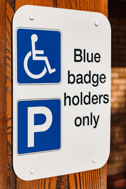 A blue parking sign indicating accessible parking with a white wheelchair symbol and an arrow pointing to the right, mounted on a metal pole in a landscaped outdoor area. The background shows blurred cars parked in the street, green bushes, and hints of buildings with warm lighting at dusk. The image highlights the designated accessible parking space outside a property, relevant to house removals and relocation services, with the sign situated on a paved pavement next to well-maintained greenery. The natural lighting and environment suggest a residential or urban setting, supporting the context of assisting clients with accessible parking when planning a home relocation or furniture transport by Man with Van Honor Oak.