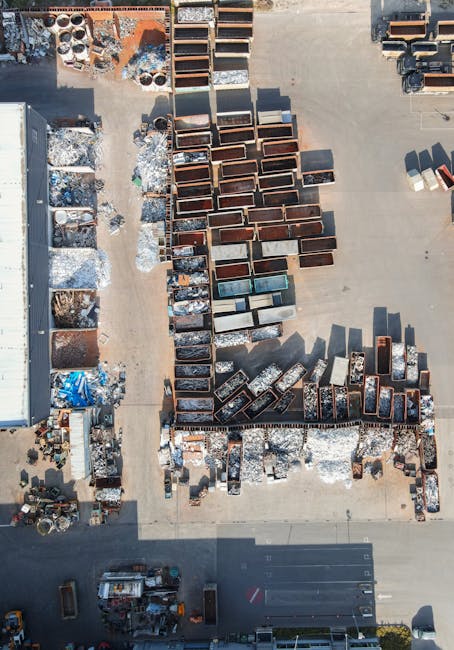 An aerial view of a large outdoor storage yard containing rows of metal and wooden furniture, including chests, cabinets, and shelves, arranged on concrete ground. Some items are stacked or placed side by side, with visible signs of use such as rust or scratches. To the left, there are scattered packing materials like plastic sheets and cardboard boxes. A narrow pathway runs through the middle of the yard, separating different sections of furniture. Around the edges, there are additional items such as large rolls of packing material and smaller pieces. The scene is well-lit by natural daylight, and in the lower part of the image, part of a vehicle with a white body is visible. This setting represents a typical furniture storage or packing area used during house removals and home relocation services, illustrating the preparation and organization involved in furniture transport by [COMPANY_NAME].