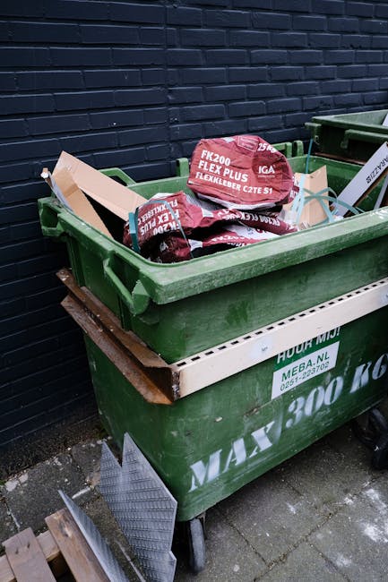 A green wheeled waste bin filled with several red and black bags of construction or garden waste, positioned outdoors on a concrete pavement against a dark brick wall. Some cardboard packaging and plastic sheeting are visible atop the waste bags, with a white construction debris container underneath. The bins are part of a home relocation or clearing process, with visible straps and handles indicating preparation for transport or disposal, typical of furniture transport or packing and moving activities associated with house removals. The area is lit evenly, highlighting the contents and surroundings, supporting a professional moving and waste management operation by Man with Van Honor Oak.