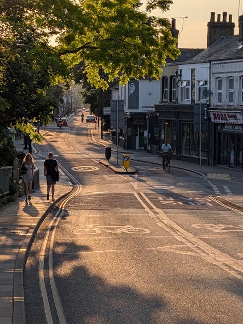 A quiet street scene in Honor Oak during late afternoon or early evening, with sunlight casting long shadows across the tarmac road. The street is lined with traditional terraced houses featuring shopfronts on the right side, including a pub or café with signage. Pedestrians, including a man and woman walking together and another cyclist, are visible on the pavement and cycle lane. A tree with lush green foliage overhangs the street, creating dappled light and shade on the road. Vehicles are seen in the background, parked along the curb and moving in the distance. The scene captures a peaceful moment in a residential and commercial area, suitable for house removals or home relocation logistics conducted by companies like Man with Van Honor Oak.