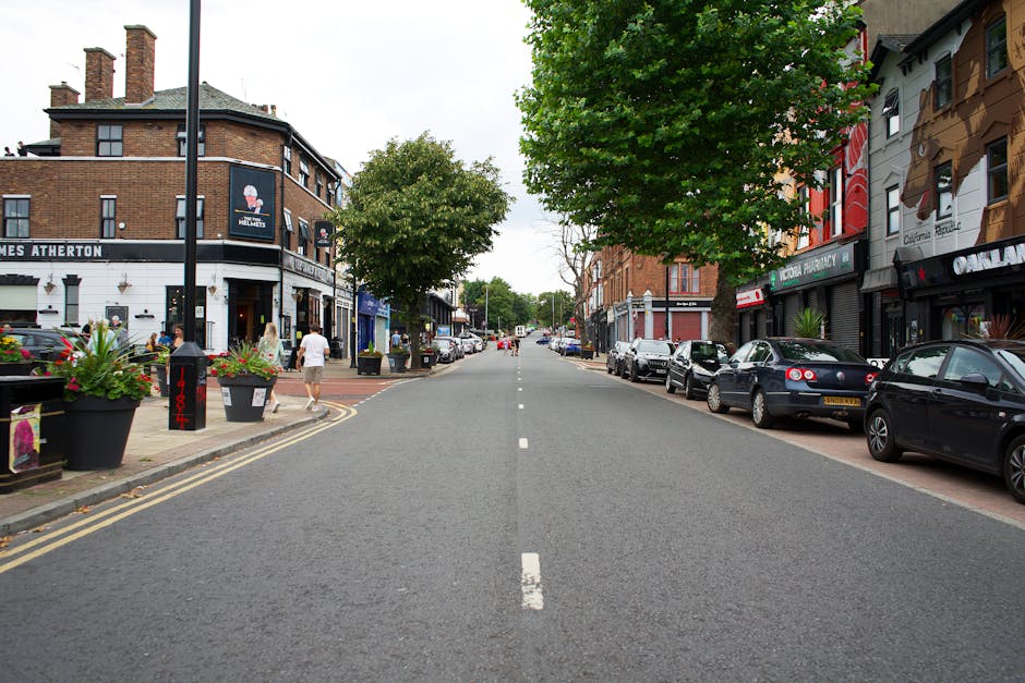 A photo of a quiet urban street in Honor Oak, featuring a row of parked cars on the right side alongside commercial shops and residential buildings. On the left side, there are pedestrians walking on the sidewalk near a building with signage and large planters filled with colorful flowers. In the background, there are trees lining the street, which is paved with asphalt and has a central dashed white line. The scene appears to be during daytime with natural light. In the context of home relocation, this image represents an exterior view where furniture and boxes might be loaded onto a van managed by Man with Van Honor Oak, as part of packing and moving processes during an urgent move-out.