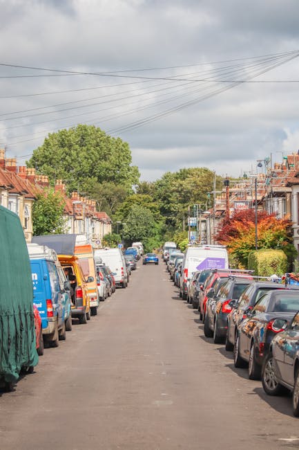 A residential street scene with a narrow asphalt road lined on both sides by parked vehicles including vans, cars, and delivery trucks, some of which are covered with protective tarps and packaging materials. The image shows a typical home relocation environment with a variety of furniture and boxes, some visible near the vehicles, indicating packing and moving preparations. In the background, there are trees and a partly cloudy sky overhead, with overhead power lines crossing the scene. The street features terraced houses with red brick facades, small front gardens, and some greenery. At the front, a white van belonging to Man with Van Honor Oak is partially visible, suggesting the presence of a professional removals service engaged in furniture transport or loading activities, aligned with the context of addressing house removal challenges such as lane blockages. The overall scene captures the logistical complexity of home relocations in urban areas, emphasizing loading and parking, which are typical considerations for professional movers.
